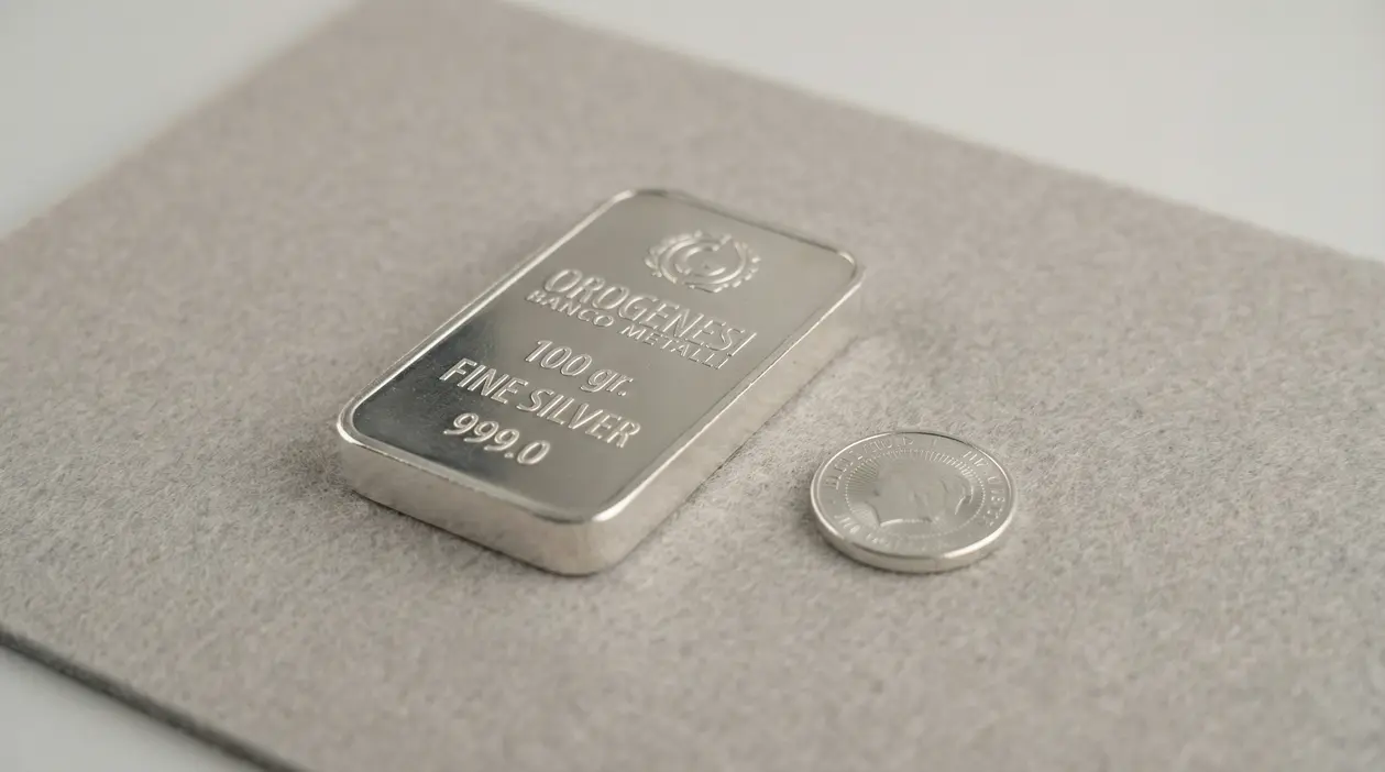 Lingotto d'argento da 100 grammi accanto a una moneta su una superficie chiara